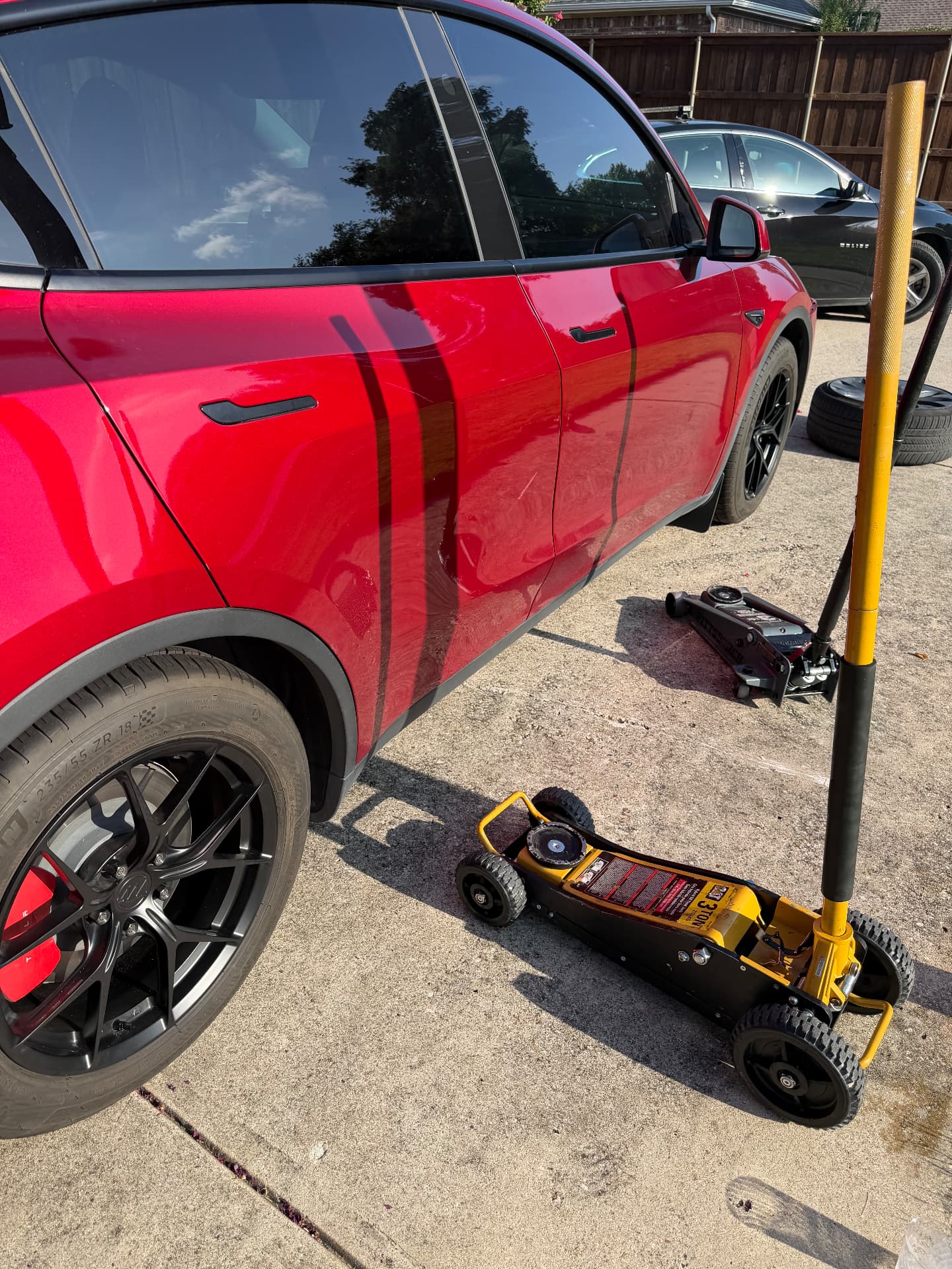Two low-profile floor jacks and a breaker bar positioned at the factory lift point of a red Tesla Model Y during a driveway wheel replacement