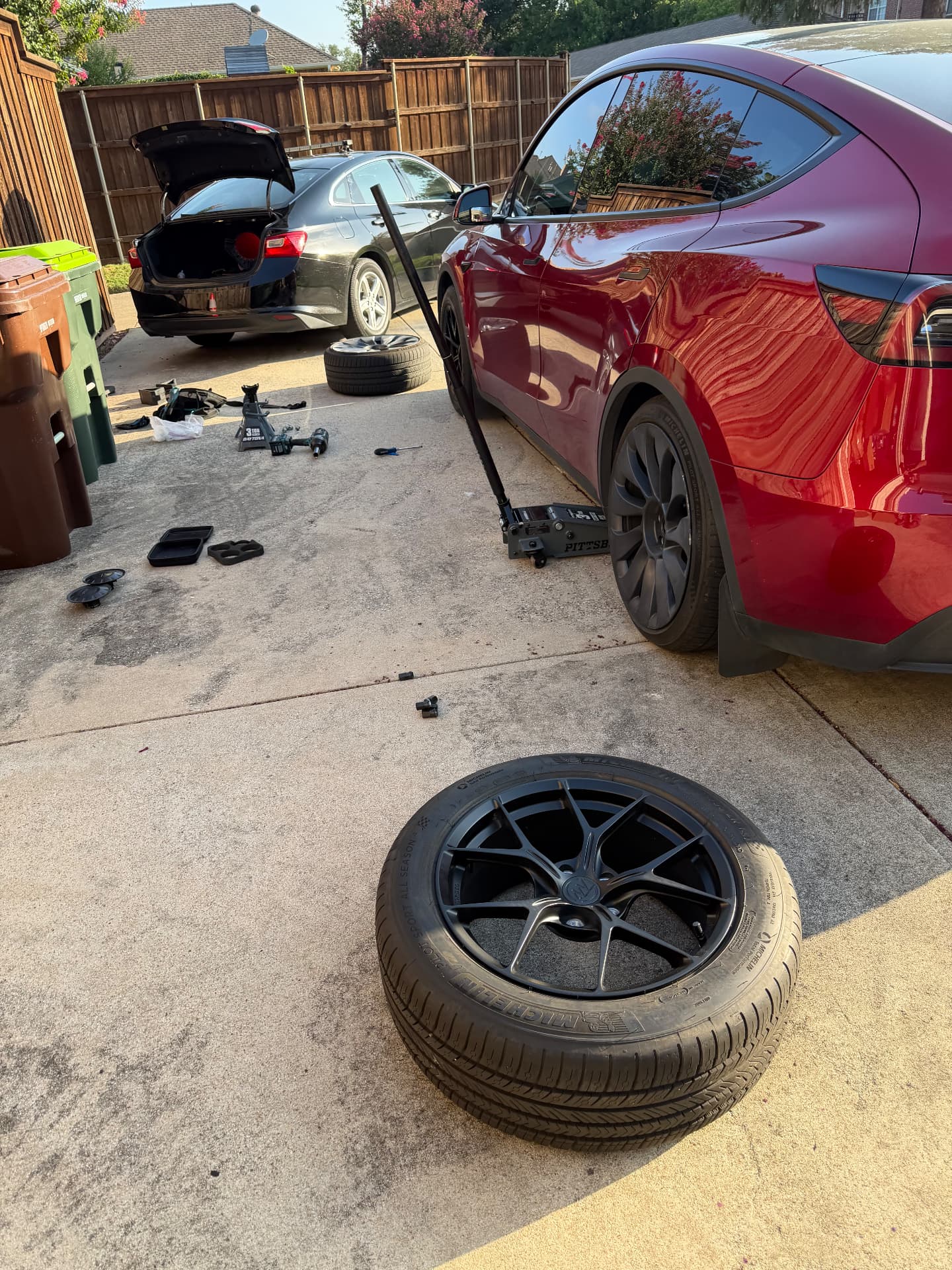 Tesla Model Y raised on an EV-rated floor jack while the damaged Michelin 235/55 ZR18 wheel sits on the driveway during a mobile tire change in Dallas–Fort Worth