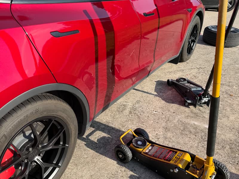 Two low-profile floor jacks and a breaker bar positioned at the factory lift point of a red Tesla Model Y during a driveway wheel replacement